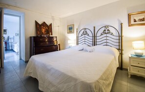 Una cama bien hecha con cabecera de metal, un aparador de madera y una mesita de noche con lámpara.