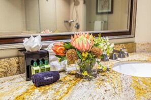A bathroom vanity with a mirror, a sink, toiletries, and a decorative flower arrangement. - Honua Kai | 4 FREE CARS | KBM Resorts | Wrap Around Lanai | 4 Units, 9 Bedrooms | HK ML-3371 (Lahaina)