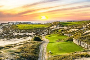 Een golfbaan met een zandpad, groene fairways en een ondergaande zon op de achtergrond.