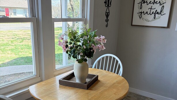 Et spiseområde med et rundt træbord, hvide stole, en vase med blomster og udsigt gennem store vinduer.