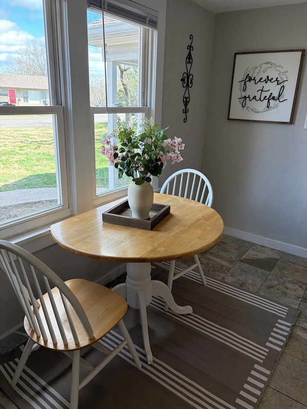 A dining area with a round wooden table, white chairs, a vase with flowers, and a view through large windows.