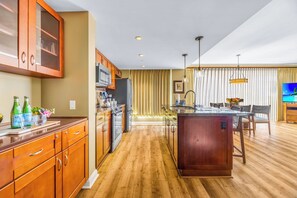 A modern kitchen with wooden cabinets, a central island, and a dining area with a TV. - Honua Kai | Ocean View 9 BR, Sleeps 26 | Car Incl. w/ 6+ Nights | HKK ML-3386 by KBM (Lahaina)