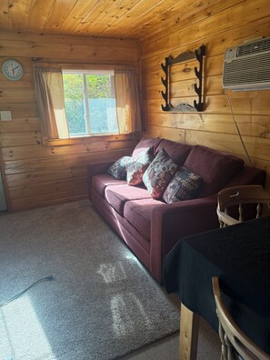 Interior de una cabaña de madera con un sofá, una ventana con cortinas, un reloj de pared y un aire acondicionado instalado en la pared.