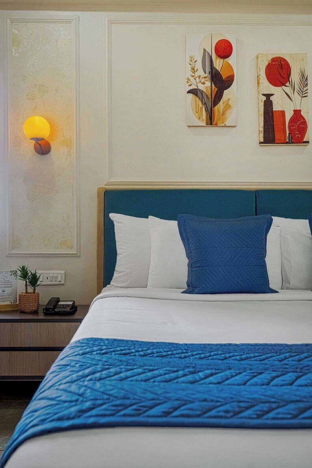 Een hotelkamer met een bed, een nachtkastje, een lamp en twee kunstwerken aan de muur.