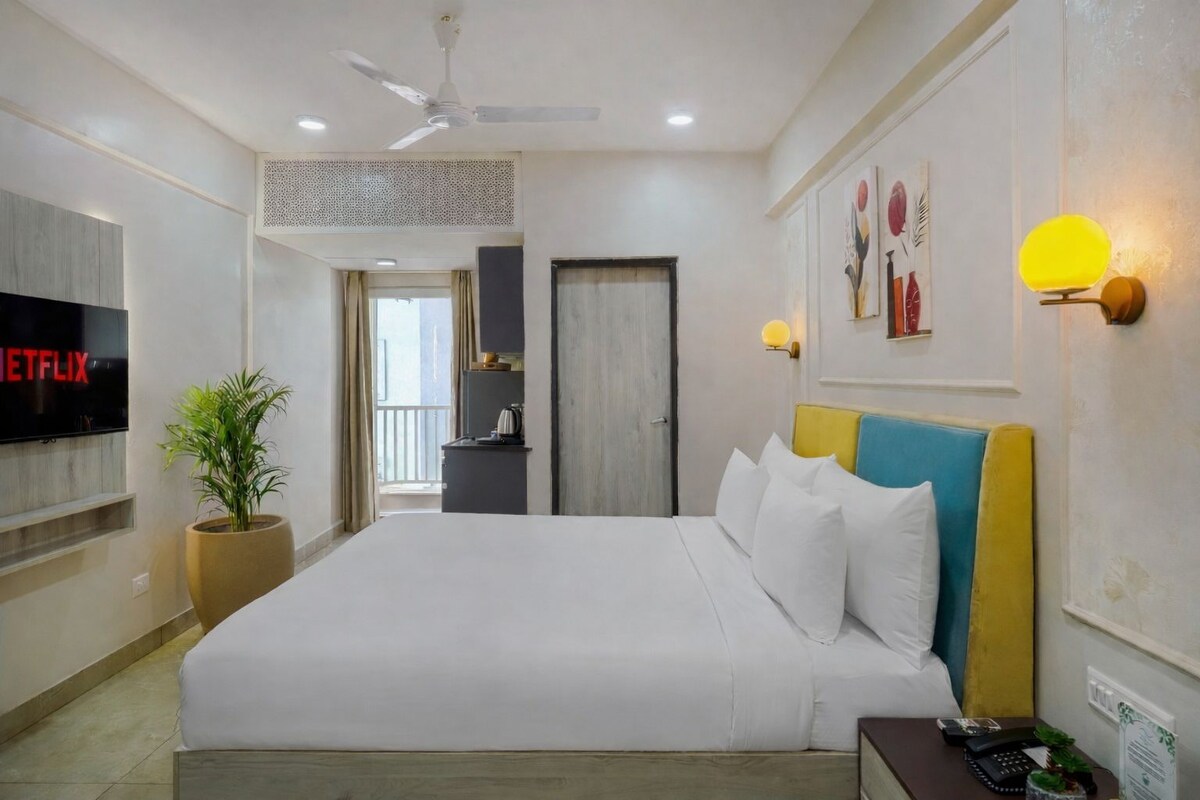 A hotel room with a bed, a TV displaying 'NETFLIX,' a potted plant, a desk with a phone, and a door leading to another room.