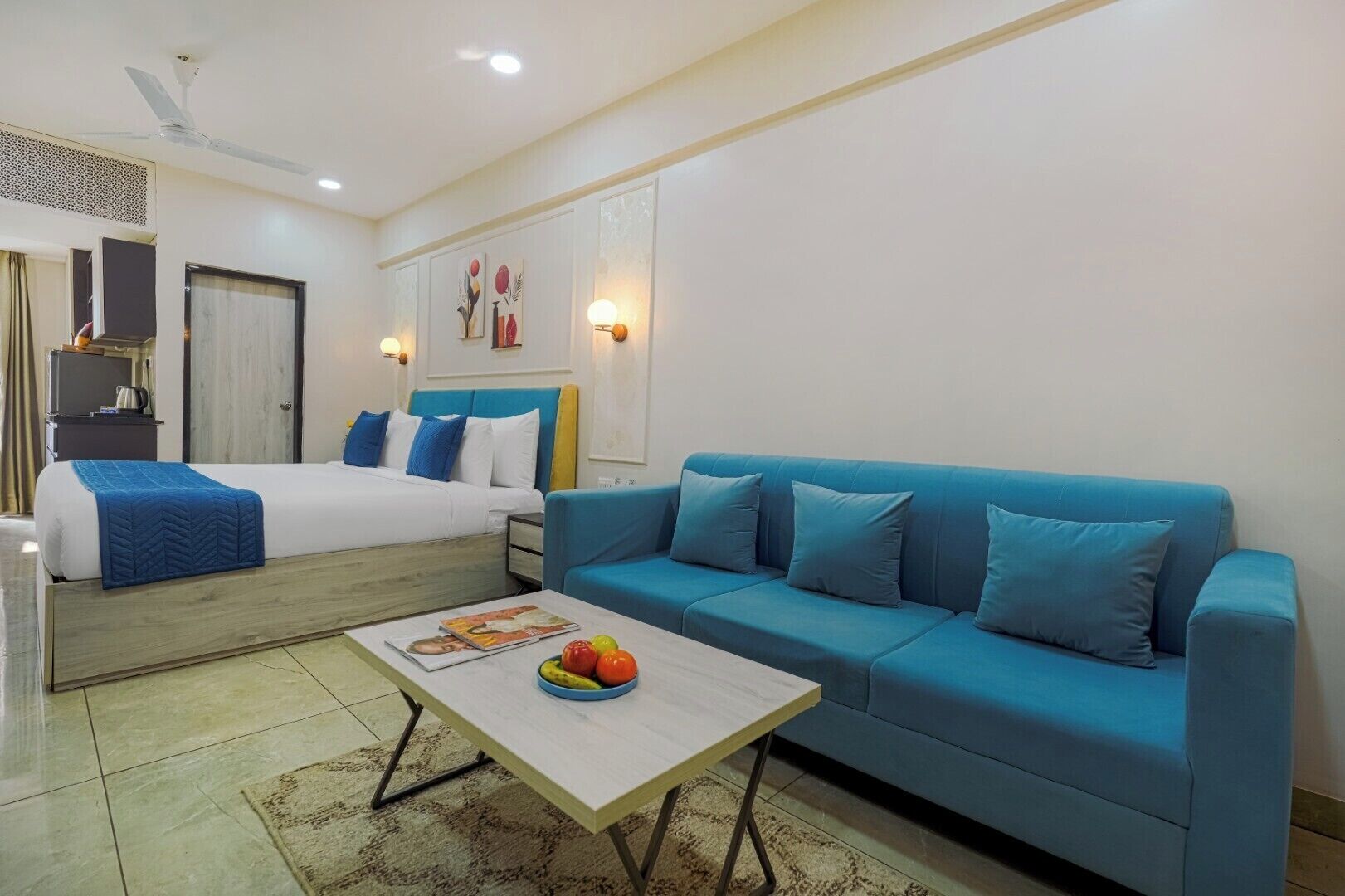 Habitación de hotel con una cama, un sofá azul, una pequeña mesa con fruta y un ventilador de techo.