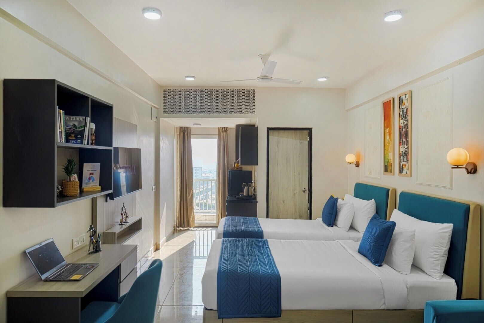 A hotel room with a bed, a desk with a laptop, a bookshelf, and a view of the city.