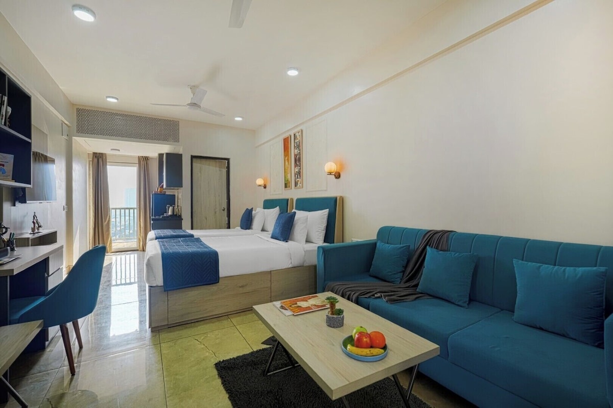 A hotel room with a bed, a blue sofa, a dining table with fruit, a desk, and a TV.