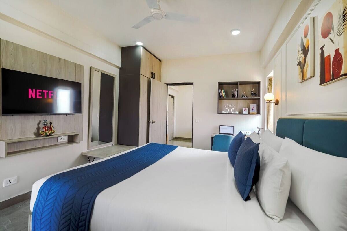 A hotel room with a bed, a TV mounted on the wall, a desk with a computer, and a bookshelf with books and decorative items.