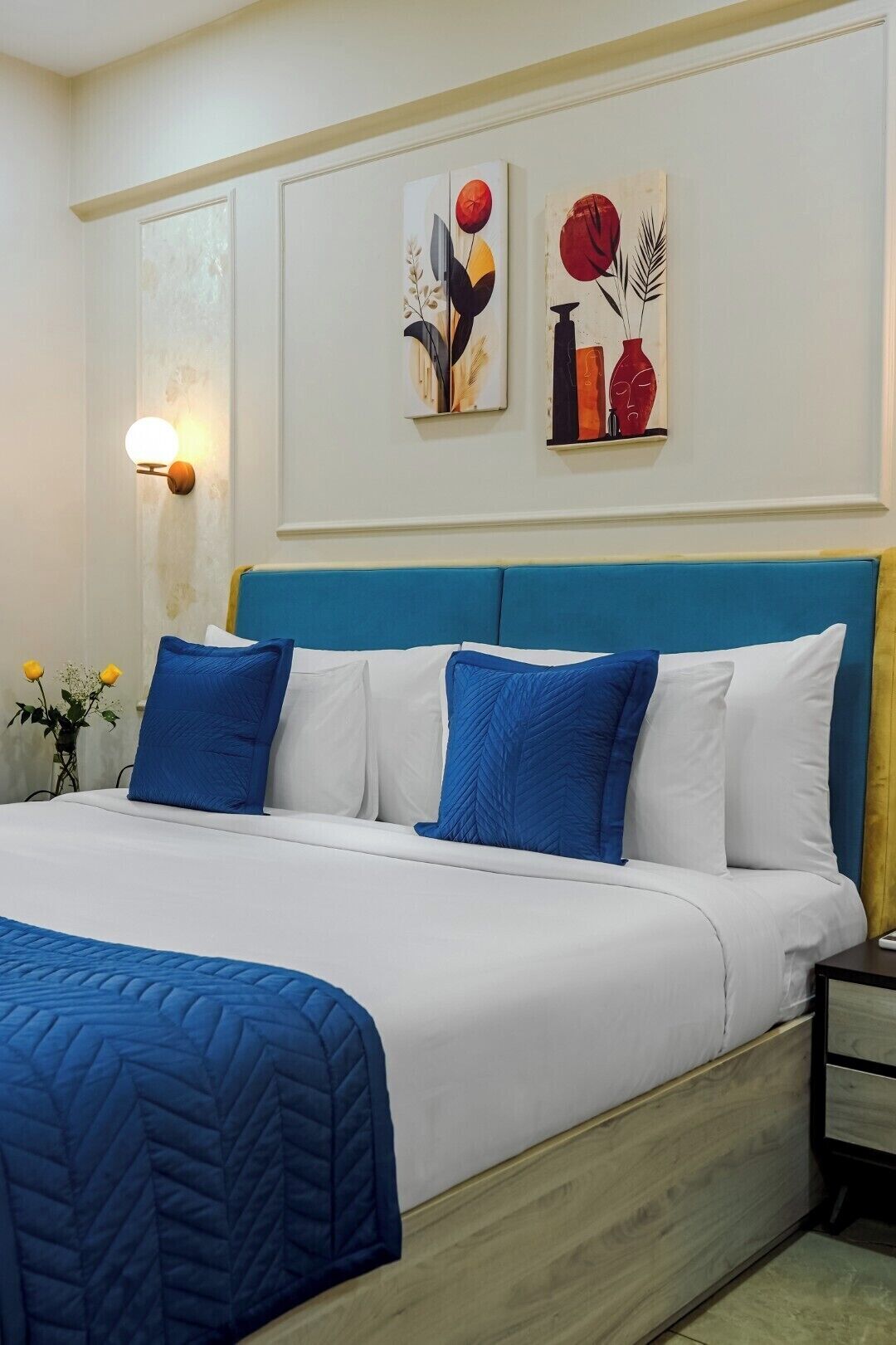 Un dormitorio con cama, ropa de cama azul y blanca, cabecera azul, mesita de noche con lámpara y dos cuadros en la pared.