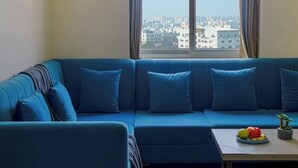 Una sala de estar con un sofá azul, una mesita con fruta y vistas a un paisaje urbano a través de una ventana.