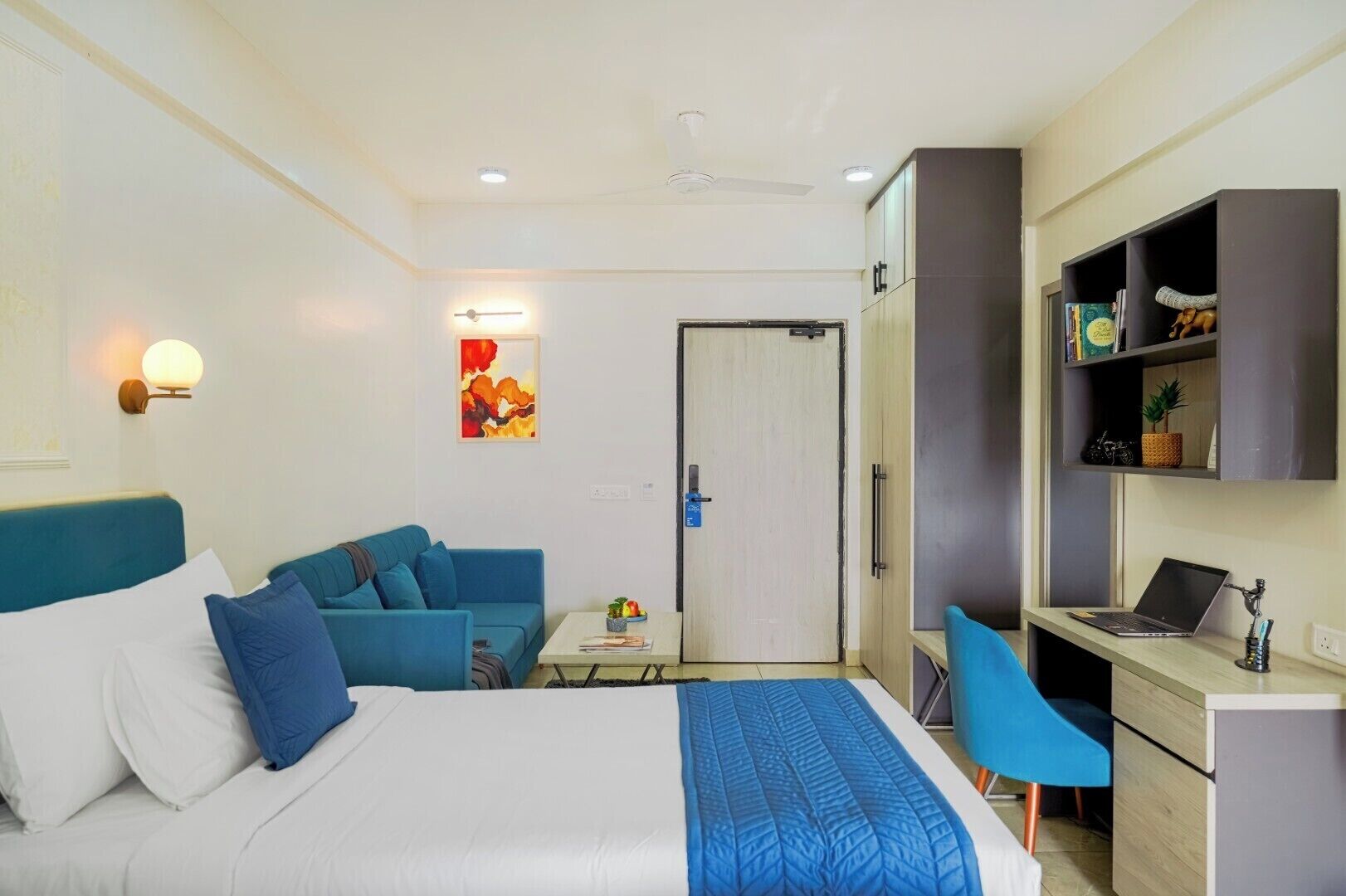 A hotel room with a bed, a sofa, a desk with a laptop, a small table with a fruit bowl, a door, and a wall-mounted shelf with decorative items.
