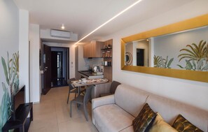 Un soggiorno moderno con un divano, un tavolo da pranzo e sedie. È presente un'area cucina con pensili e uno specchio.