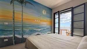 Une chambre avec une fresque représentant un coucher de soleil sur l’océan, un lit et un balcon avec vue.