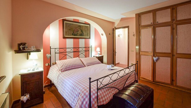 Une chambre à coucher comprenant un lit, une table de chevet, une lampe, un banc, une armoire et une porte en arc de cercle.