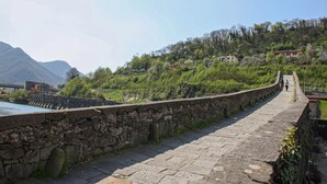 Un pont de pierre, avec un mur de pierre d’un côté et un sentier menant à une colline où se trouvent des bâtiments.
