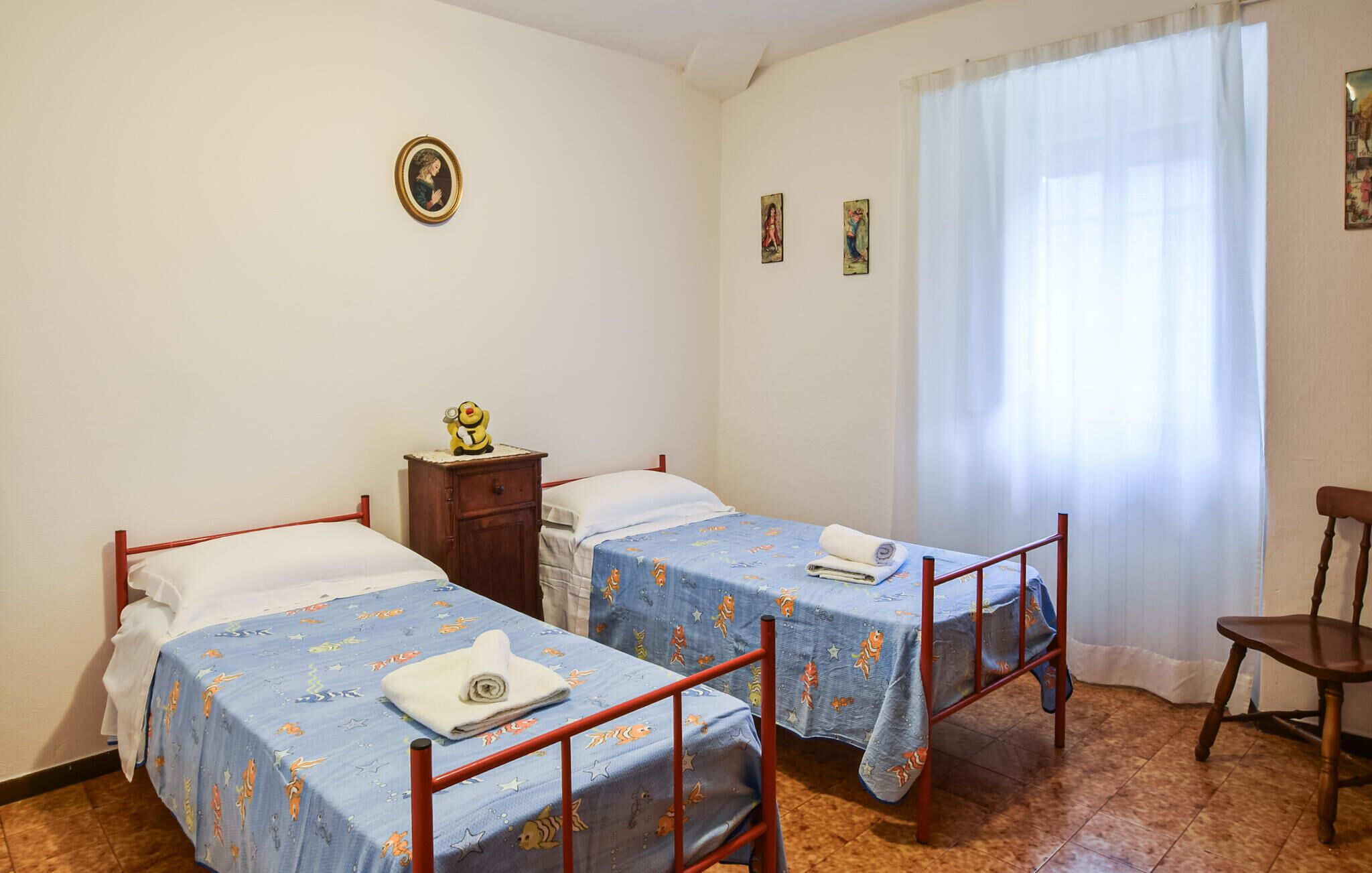 A room with two beds, a small wooden nightstand, a chair, and a window with curtains.