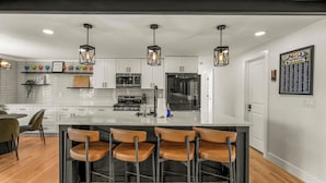 Cozinha moderna com armários brancos, uma ilha central com banquetas, iluminação pendente e um cardápio emoldurado na parede.