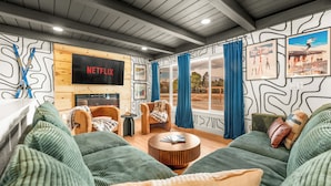Uma sala de estar moderna com um letreiro da Netflix, sofás verdes, uma mesa de centro de madeira e uma grande janela com cortinas.