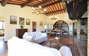 Een ruime woonkamer met een hoge plafond, een eethoek met een grote houten tafel, en een keuken met een bakstenen muur en houten balken.