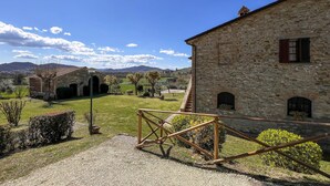 A stone building with a staircase leading to a door, surrounded by a grassy area with a metal gate. - Umbria Magione Podere C. (Magione)