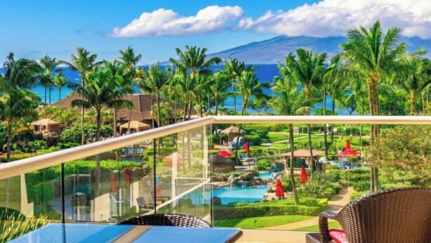 Un resort con balaustrada de vidrio, piscina al aire libre, palmeras y vistas al océano y una montaña.