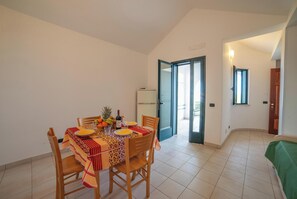 A dining area with a table set for four, chairs, a refrigerator, and a door leading to an outdoor balcony. - Beach in 5 min | Pool Access | n.66, Otranto, Italy (Otranto)
