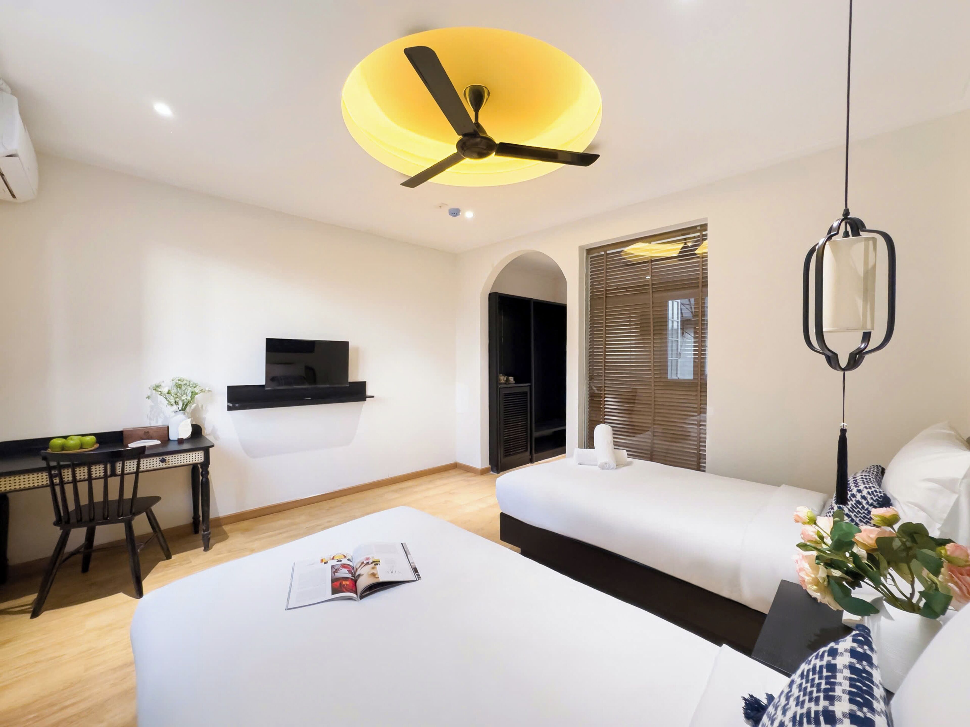 A hotel room with a bed, a desk with a TV, a ceiling fan, and a window with blinds.