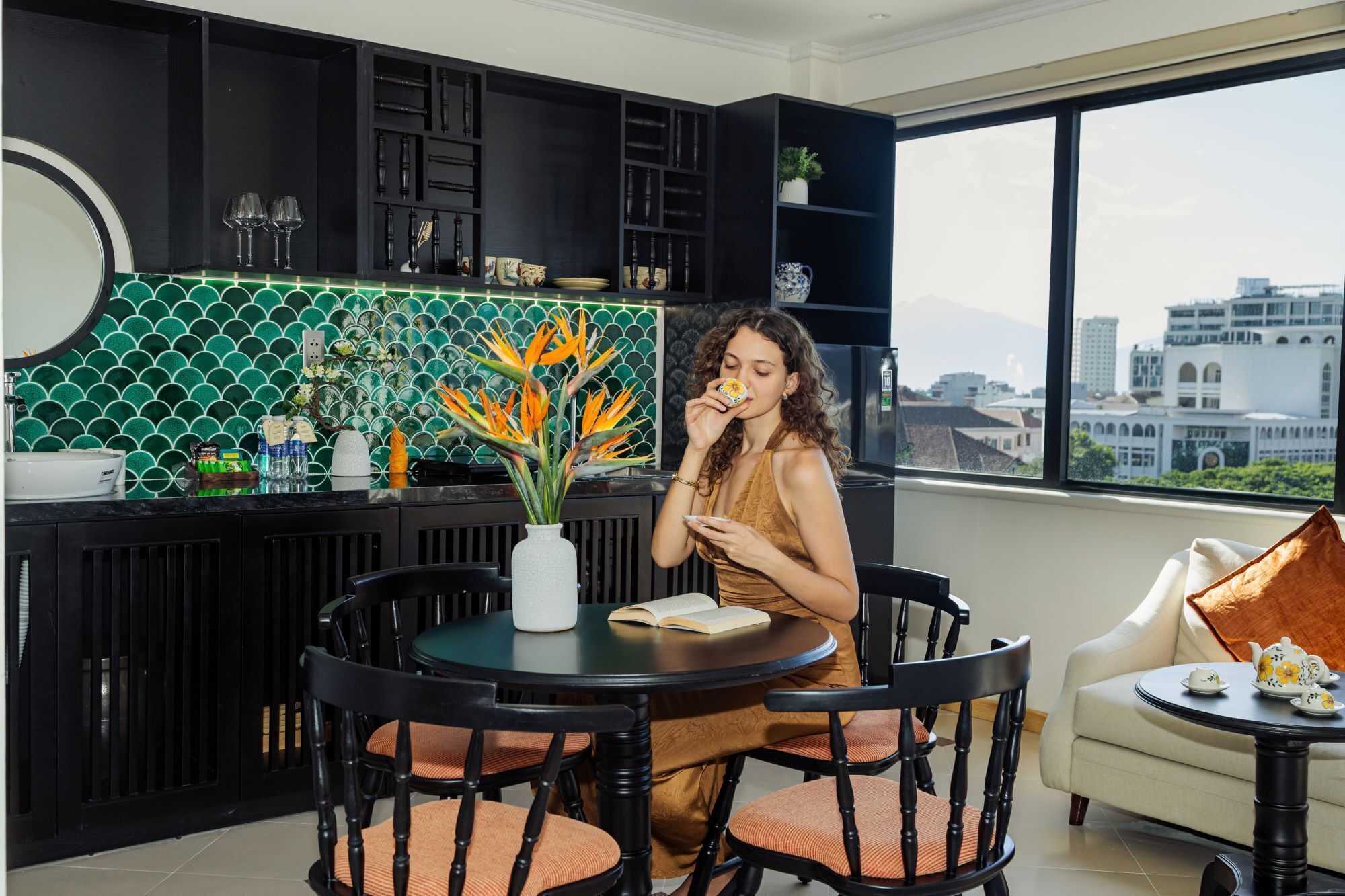 Una cocina moderna con salpicadero de azulejos verdes, una zona de comedor con una mesa redonda y sillas, y vistas a la ciudad a través de grandes ventanales.
