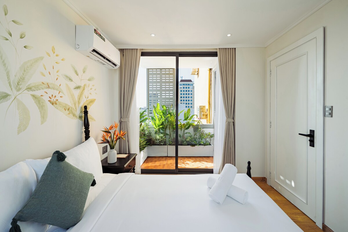 Une chambre d’hôtel avec un lit, une table de chevet, une plante en pot et une vue sur la ville, visible à travers une porte coulissante.