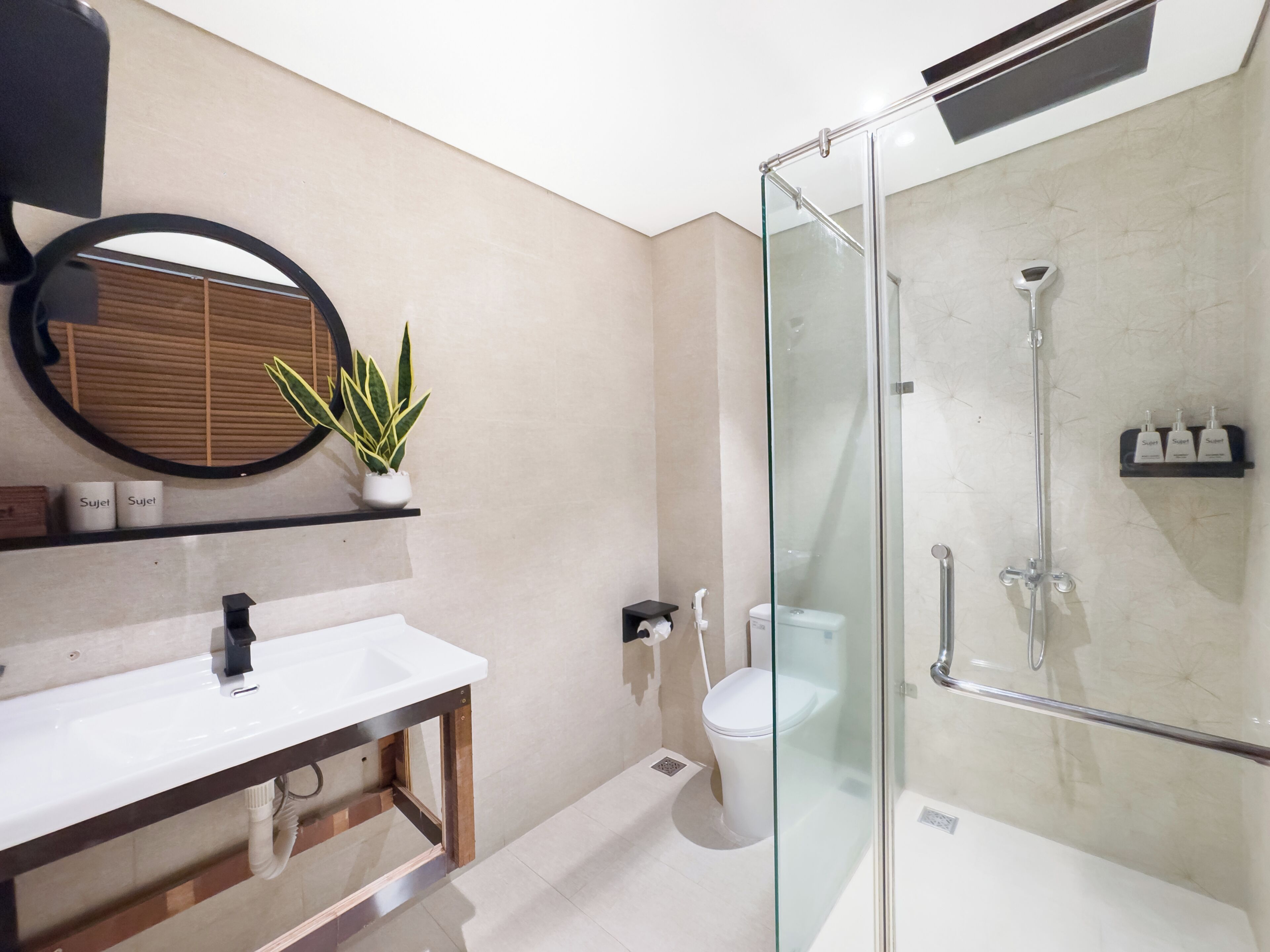 A modern bathroom with a glass shower enclosure, a round mirror, a white sink, and a potted plant.