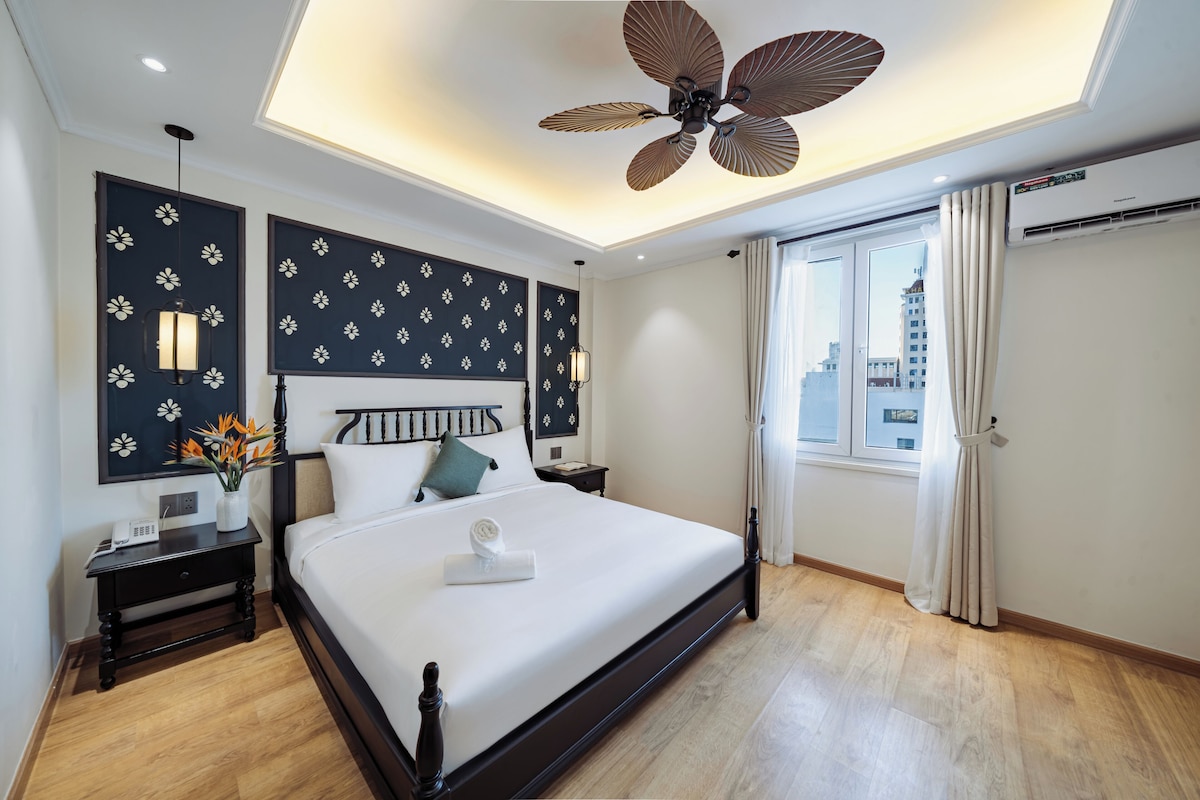 Une chambre d’hôtel comprenant un lit, une table de chevet, une lampe fixée au mur, un ventilateur de plafond et une fenêtre avec des rideaux.
