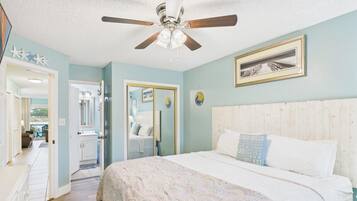 Une chambre à coucher avec un lit, un ventilateur de plafond, un tableau encadré et une salle de bain visible à travers une porte ouverte.