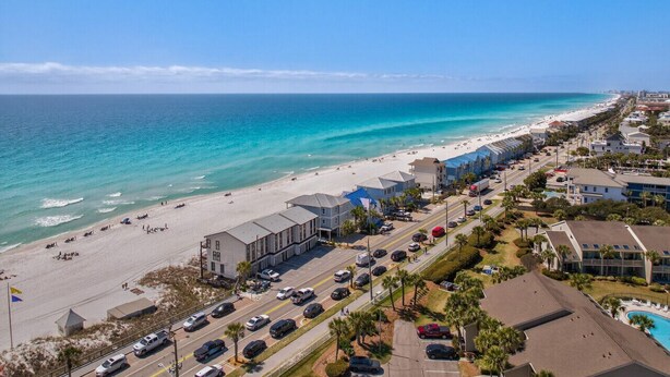 An aerial view of a coastal area with a beach, buildings, and a road.