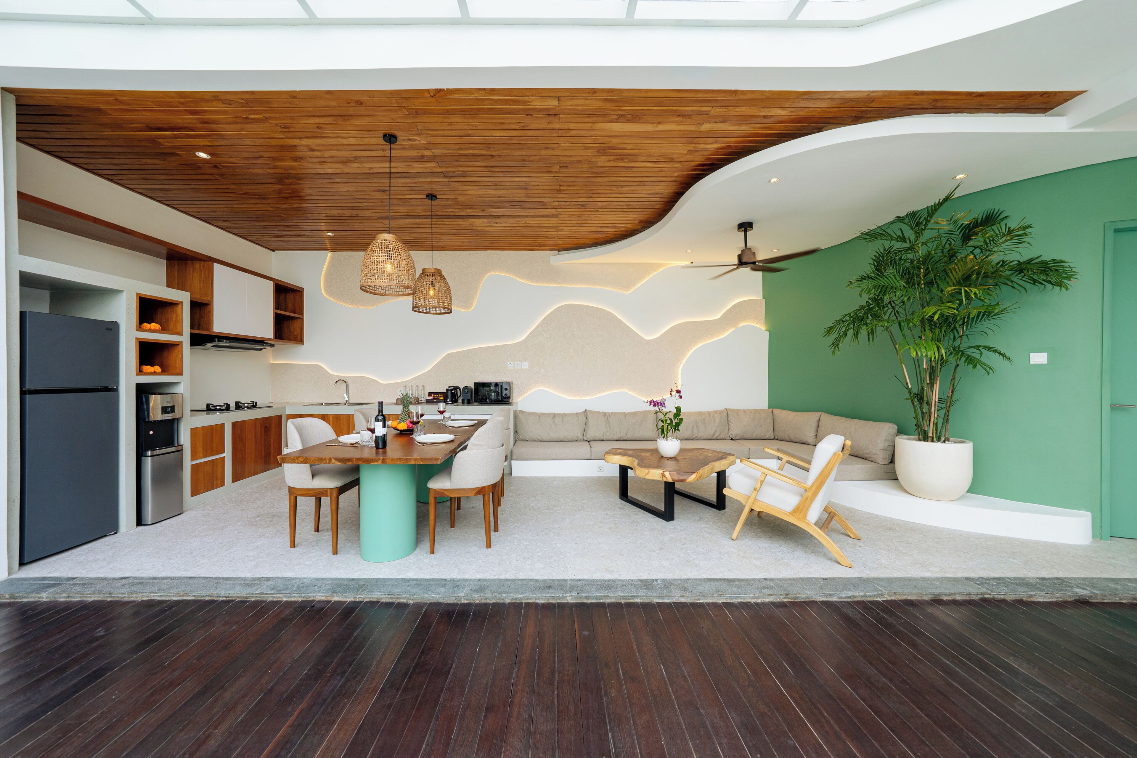 A modern kitchen with wooden cabinets, a dining area with a table and chairs, a sofa, and a large potted plant.