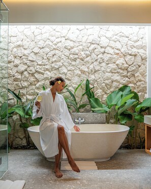 A person in a white bathrobe sitting in a bathtub with a stone wall behind them, surrounded by green plants. -  Lomo Boutique Hotel, Uluwatu. (Pecatu)