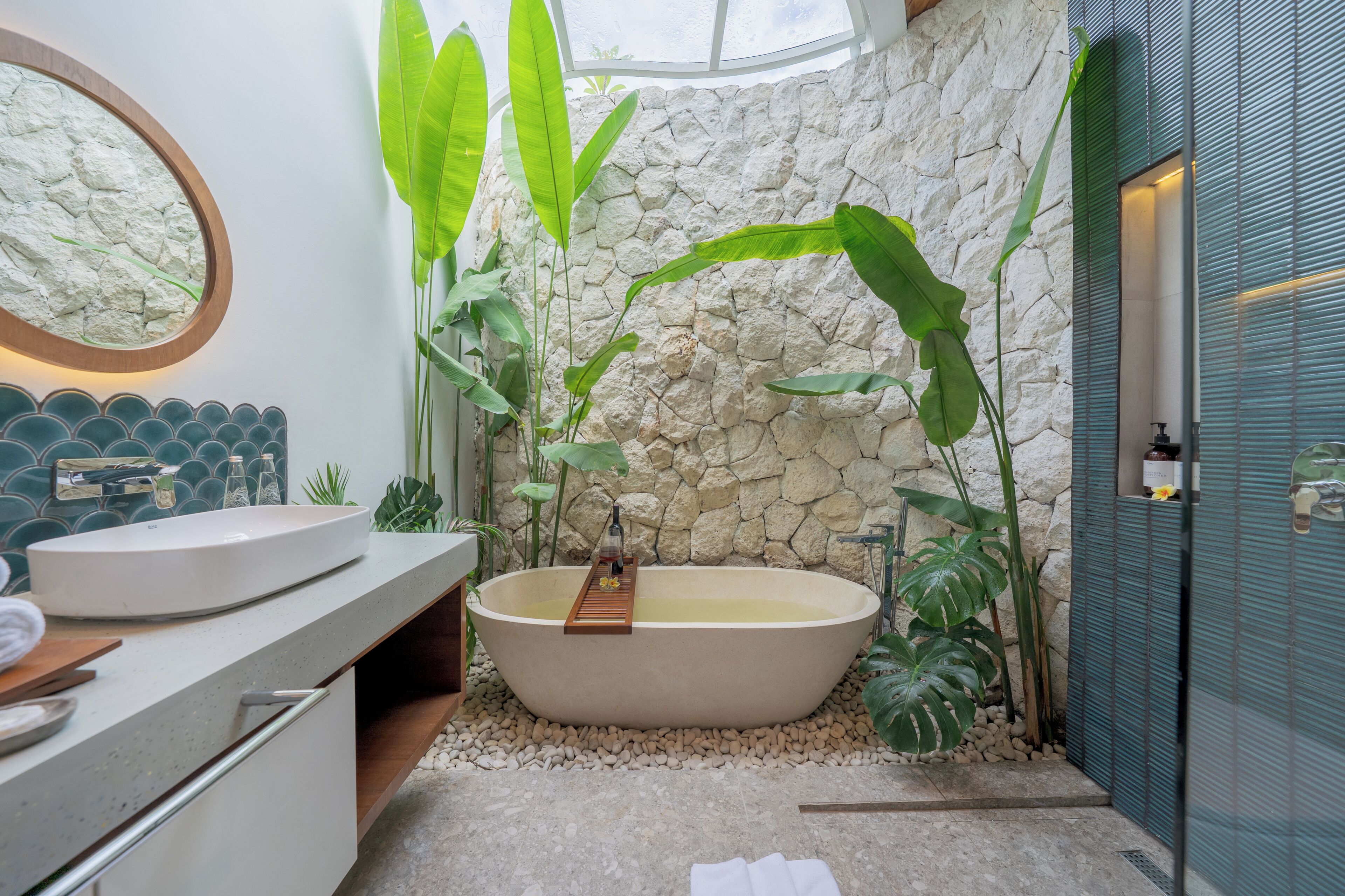 A bathroom with a freestanding bathtub, a round mirror, a sink, and a stone wall.
