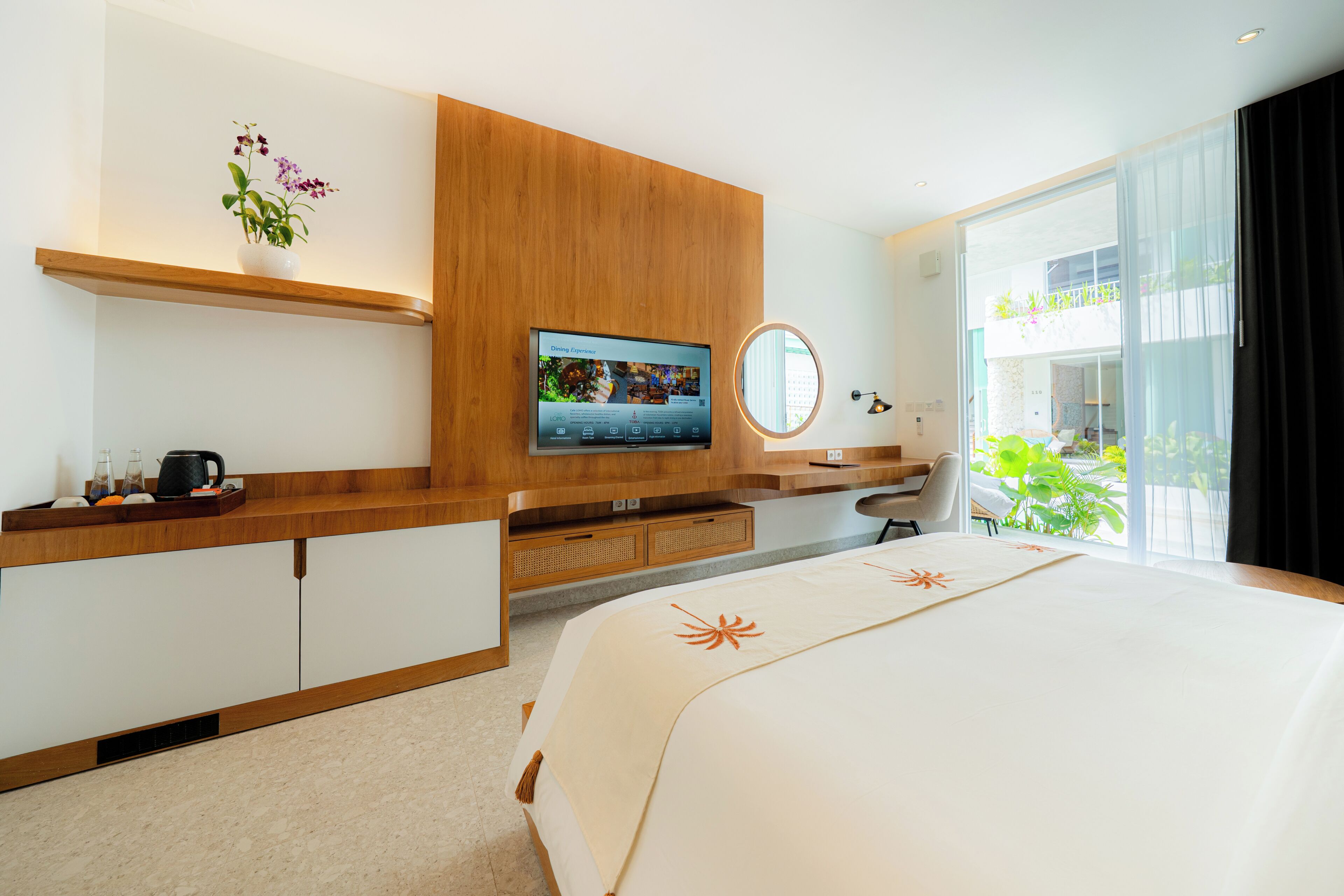 A modern hotel room with a large bed, a TV mounted on a wooden panel, a small desk, and a window with a view of greenery.