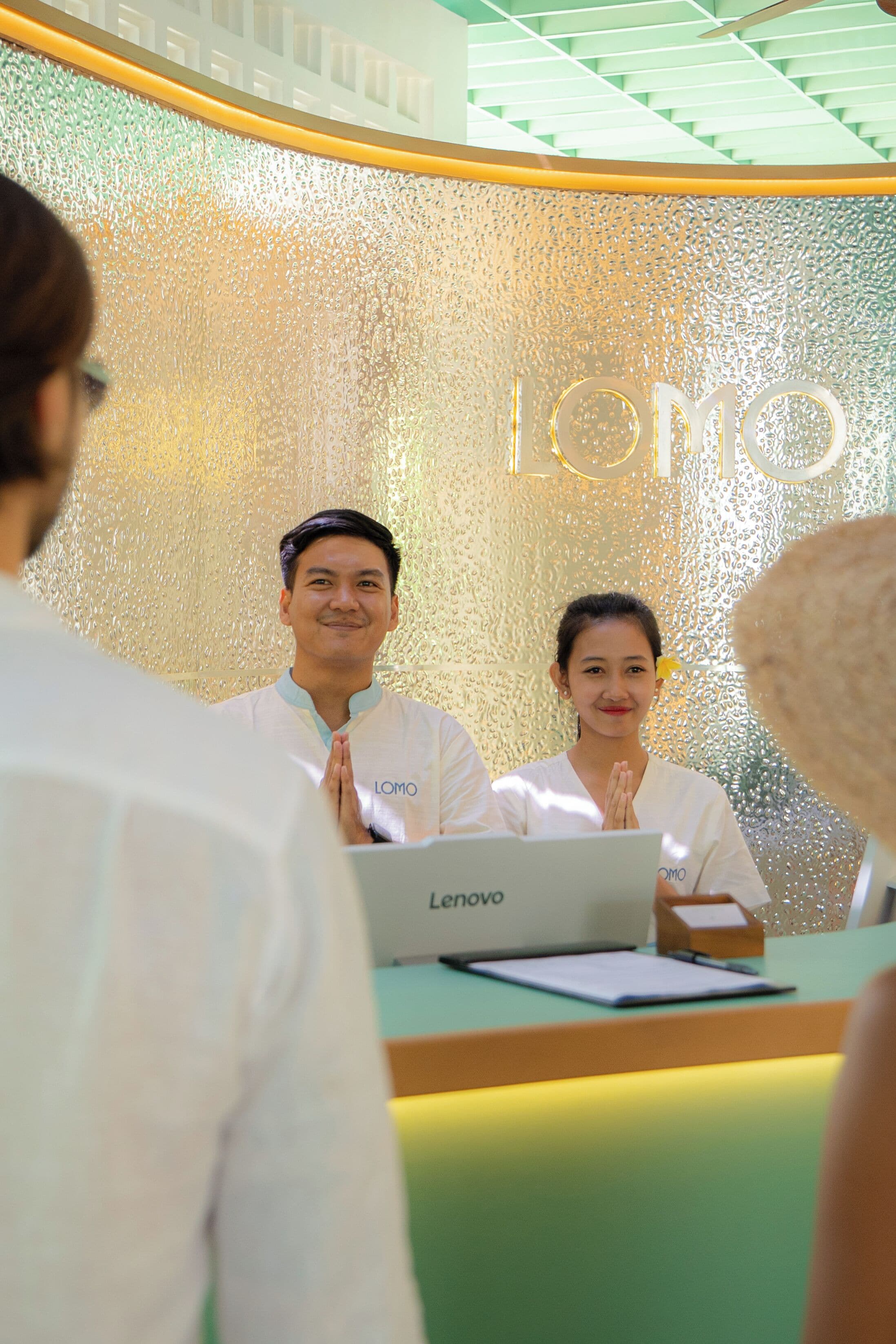 A reception desk with a Lenovo laptop, a glass wall with a textured pattern, and a sign with the word 'Lomo'.