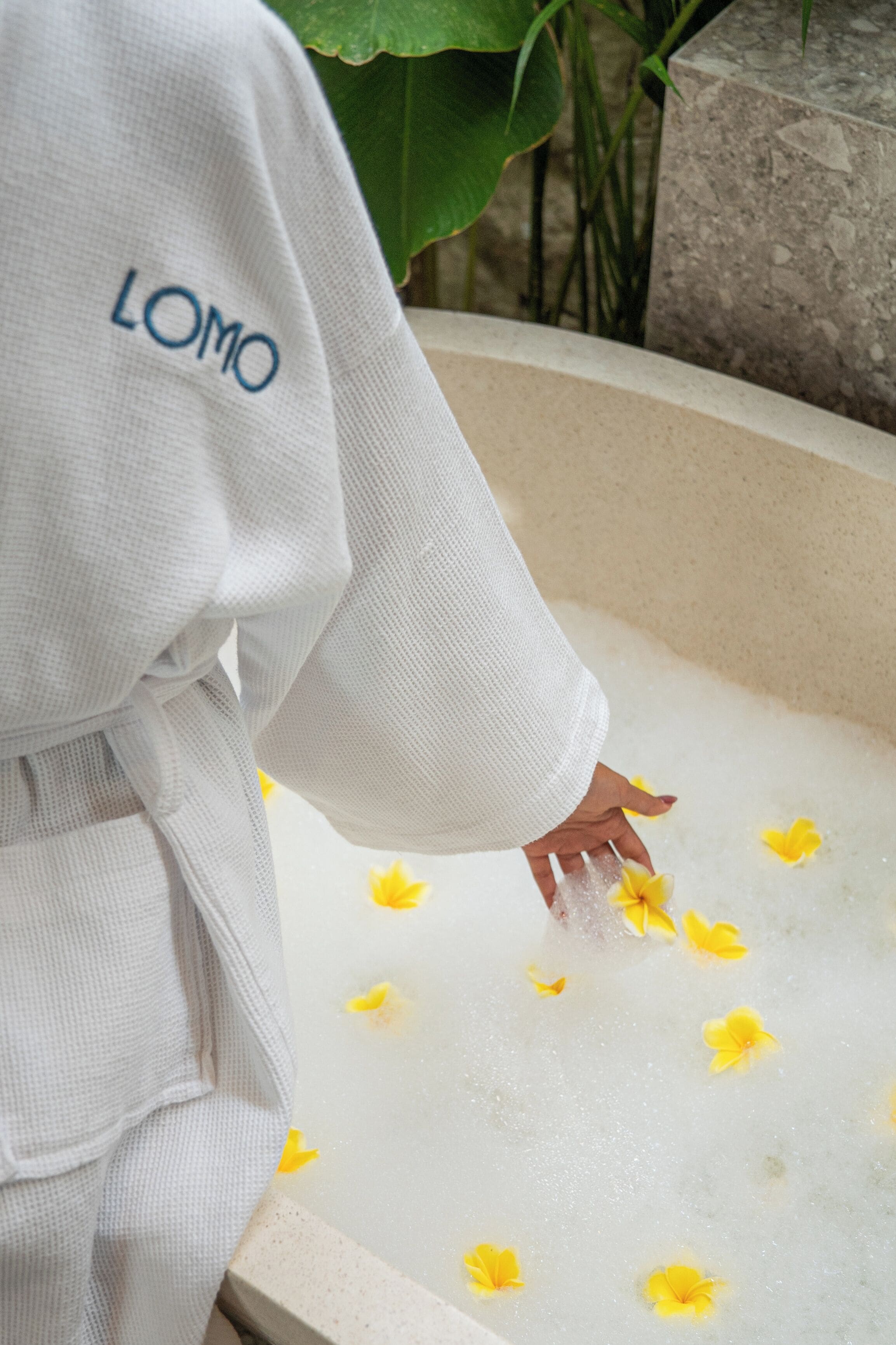 A person in a white robe with 'LOMO' text, reaching into a bathtub filled with water and yellow flowers.