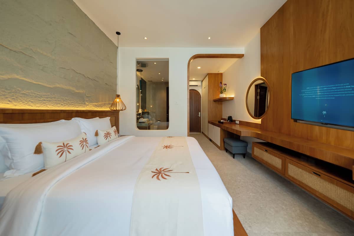 Quarto de hotel moderno com uma cama grande, televisão, secretária de madeira e casa de banho visível através de uma porta aberta.