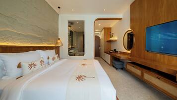 Quarto de hotel moderno com uma cama grande, televisão, secretária de madeira e casa de banho visível através de uma porta aberta.