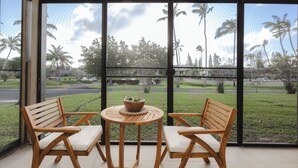 A room with two wooden chairs and a small table, large windows with a view of palm trees and a road. - Turtle Bay Renovated Luxury Condo • Walk to Beach • King Bed • AC •  (Kahuku)