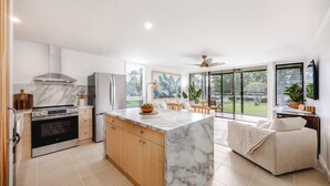 A modern kitchen with a marble island, wooden cabinets, and a large window overlooking a garden. - Turtle Bay Renovated Luxury Condo • Walk to Beach • King Bed • AC •  (Kahuku)