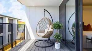 Un balcone con una poltrona a dondolo, una piccola pianta in vaso e uno scorcio di una camera da letto con un letto e una finestra.