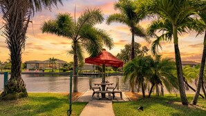 A waterfront patio with a table and chairs, surrounded by palm trees and a body of water. - Waterfront Oasis - Fish from Backyard, Heated Pool & Spa (Cape Coral)