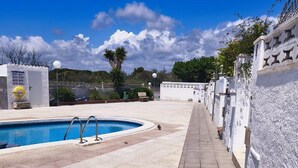 Una piscina con borde de baldosas, una pared blanca con detalles decorativos y un cielo azul despejado.