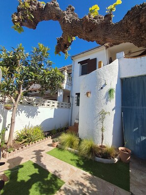 Un patio con un árbol, plantas en macetas y un edificio blanco con balcón.