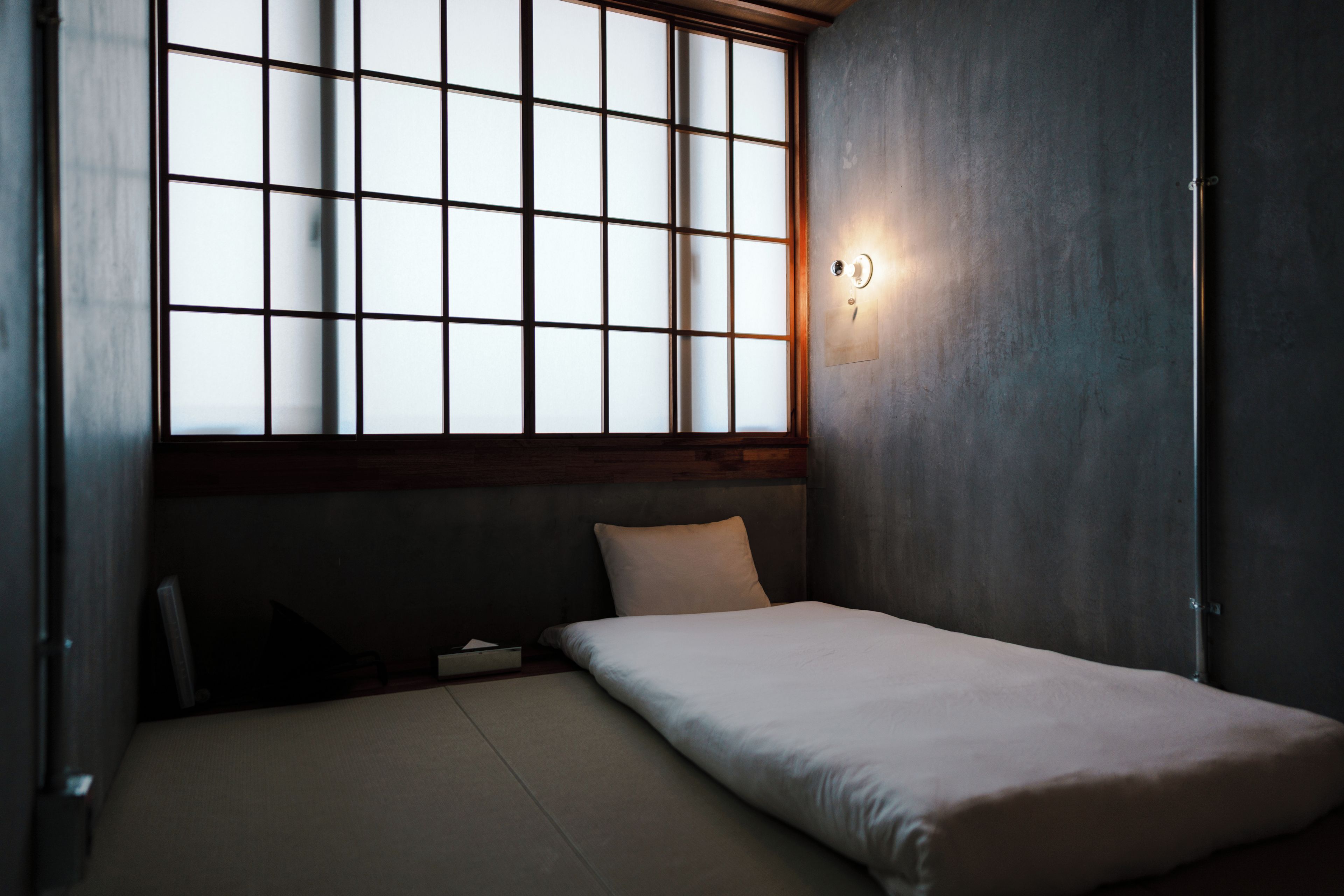 A room with a bed, a window with a grid pattern, and a wall-mounted light fixture.