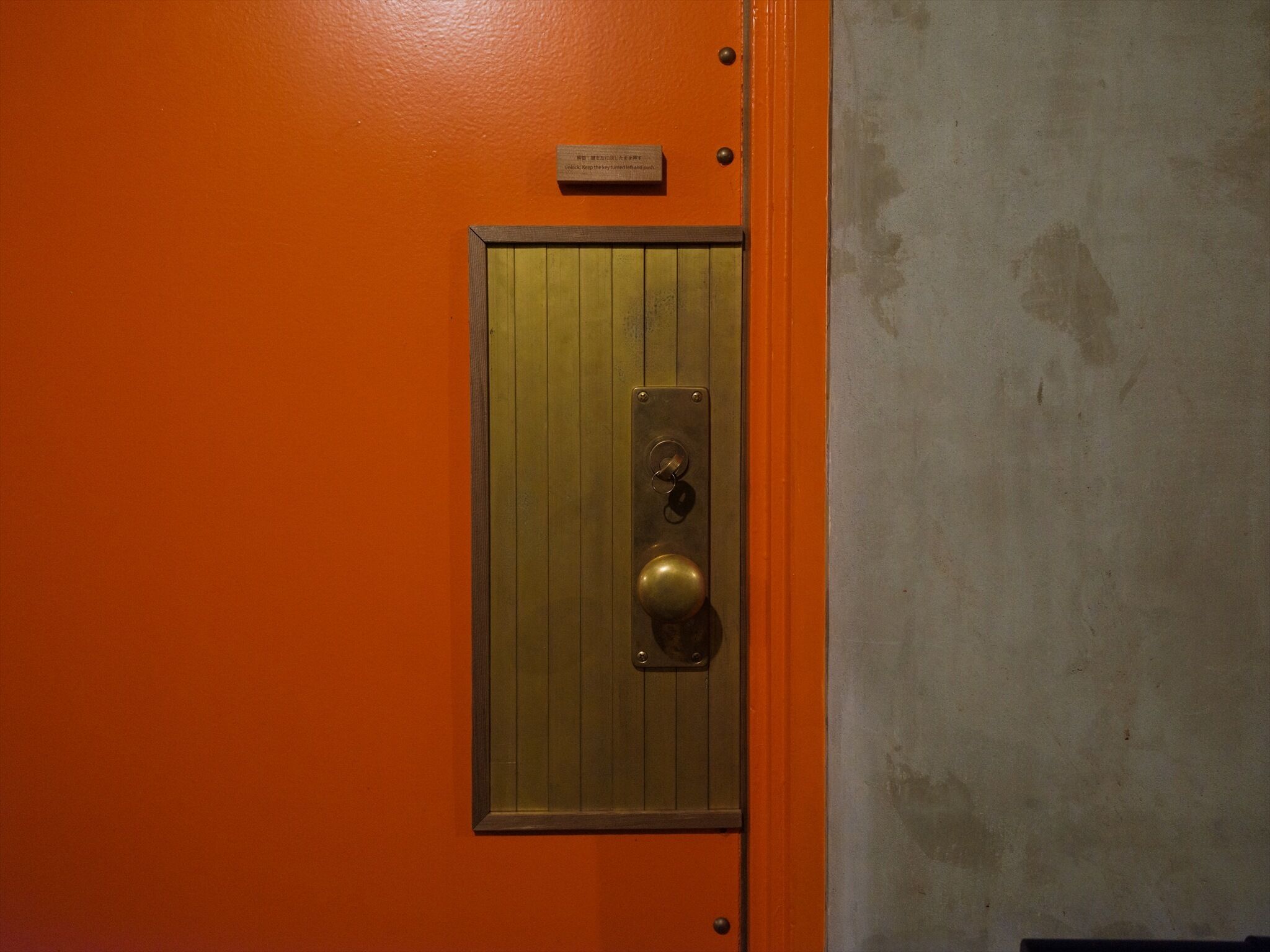 A door with a brass handle and a lock, set in an orange wall with a metal plate above it.